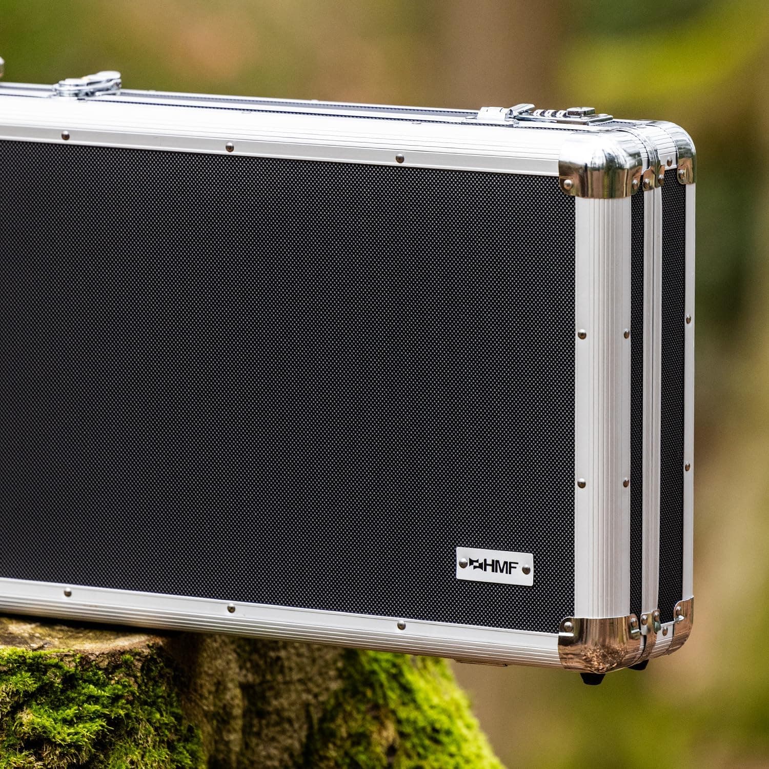 Ein abschließbarer Waffenkoffer für Gewehre, Pistolen, Airsoft oder Luftgewehr in Silber und Schwarz mit Metallkanten und kleinem HMF-Logo steht auf einem moosigen Baumstumpf, im Hintergrund unscharfes Grün.