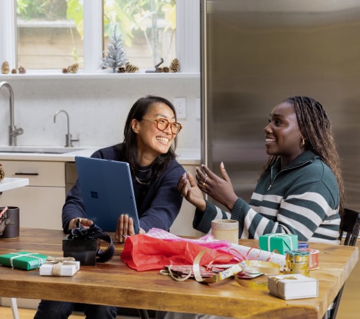Zwei Frauen sitzen lächelnd und unterhalten sich an einem mit Geschenkpapier bedeckten Küchentisch. Die eine hält einen Laptop in der Hand, die andere gestikuliert fröhlich. Verpackte Geschenke und Bänder liegen verstreut auf dem Tisch.